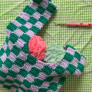 May include: A handmade crochet bag with a green and lavender checkerboard pattern. The bag contains two fluffy pom-poms, one pink and one green. A pink crochet hook with red polka dots rests on the green and white checkered background.