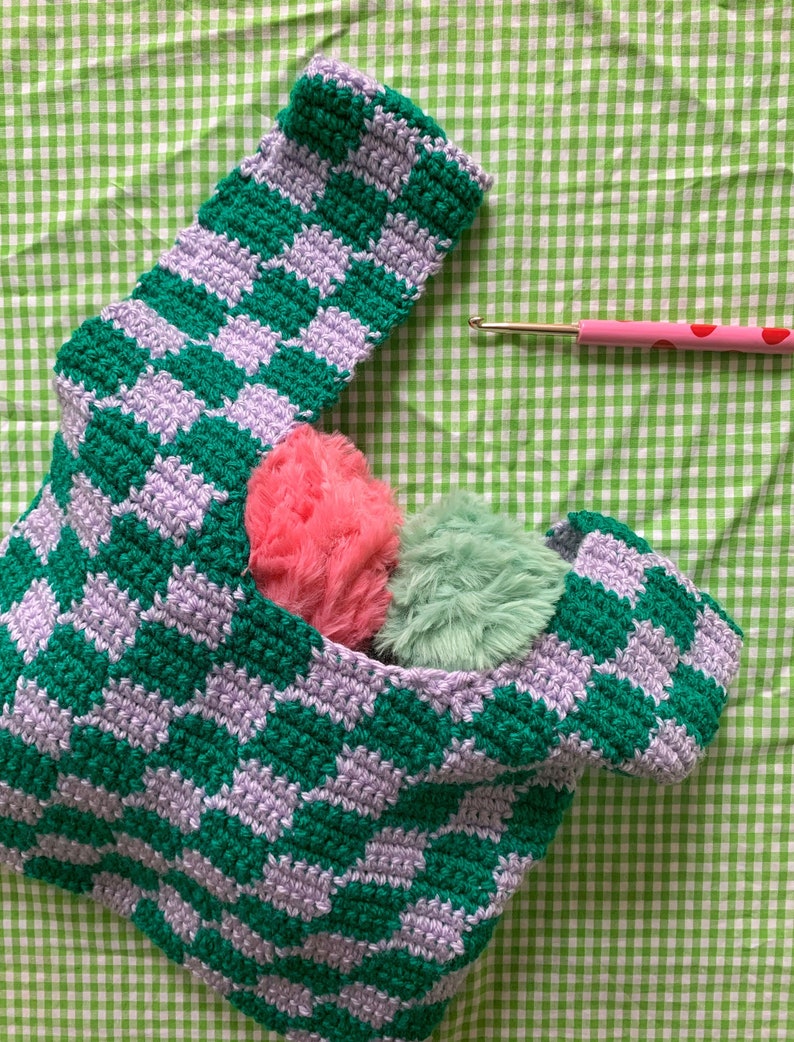 May include: A handmade crochet bag with a green and lavender checkerboard pattern. The bag contains two fluffy pom-poms, one pink and one green. A pink crochet hook with red polka dots rests on the green and white checkered background.
