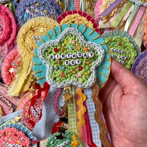 May include: A handmade crochet badge featuring a light green star with the text BIRTHDAY BOY spelled out in white letter beads. The star is layered over a turquoise felt circle and decorated with colourful sequins and gold star charms.