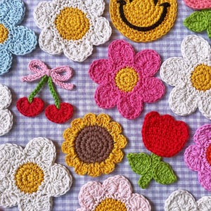 K&ouml;nnte beinhalten: Eine Sammlung farbenfroher, handgefertigter H&auml;kelartikel. Das Bild zeigt verschiedene florale Designs, darunter G&auml;nsebl&uuml;mchen, Sonnenblumen und eine rote Tulpe, zusammen mit einem gelben Smiley und einem Paar Kirschen. Die Artikel sind auf einer karierten Oberfl&auml;che angeordnet.