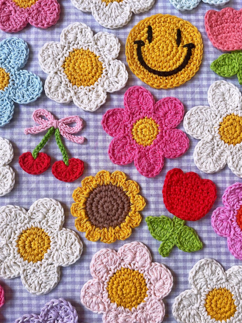 K&ouml;nnte beinhalten: Eine Sammlung farbenfroher, handgefertigter H&auml;kelartikel. Das Bild zeigt verschiedene florale Designs, darunter G&auml;nsebl&uuml;mchen, Sonnenblumen und eine rote Tulpe, zusammen mit einem gelben Smiley und einem Paar Kirschen. Die Artikel sind auf einer karierten Oberfl&auml;che angeordnet.
