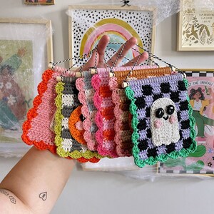May include: A collection of colorful, handmade crochet wall hangings. Each square features a unique design, including a ghost, checkerboard patterns, and scalloped edges. The hangings are in shades of pink, orange, green, and purple.