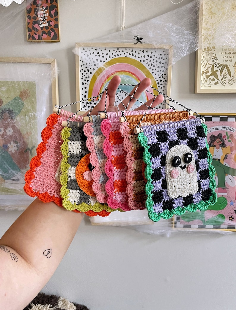 May include: A collection of colorful, handmade crochet wall hangings. Each square features a unique design, including a ghost, checkerboard patterns, and scalloped edges. The hangings are in shades of pink, orange, green, and purple.