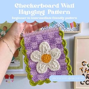 Könnte beinhalten: Ein gehäkeltes Wandbehang mit einem Schachbrettmuster in Lila, einer weißen und gelben Blume und einem grünen, gewellten Rand. Der Text "Checkerboard Wall Hanging Pattern" wird oben angezeigt.