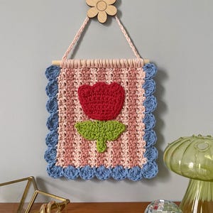K&ouml;nnte beinhalten: Handgefertigter Wandbehang aus H&auml;kelarbeit mit einer roten Blume und gr&uuml;nen Bl&auml;ttern auf einem rosa-wei&szlig; gestreiften Hintergrund. Die Kanten sind mit blauem H&auml;kelwerk verziert. Eine Holzblume dient zum Aufh&auml;ngen des St&uuml;cks.