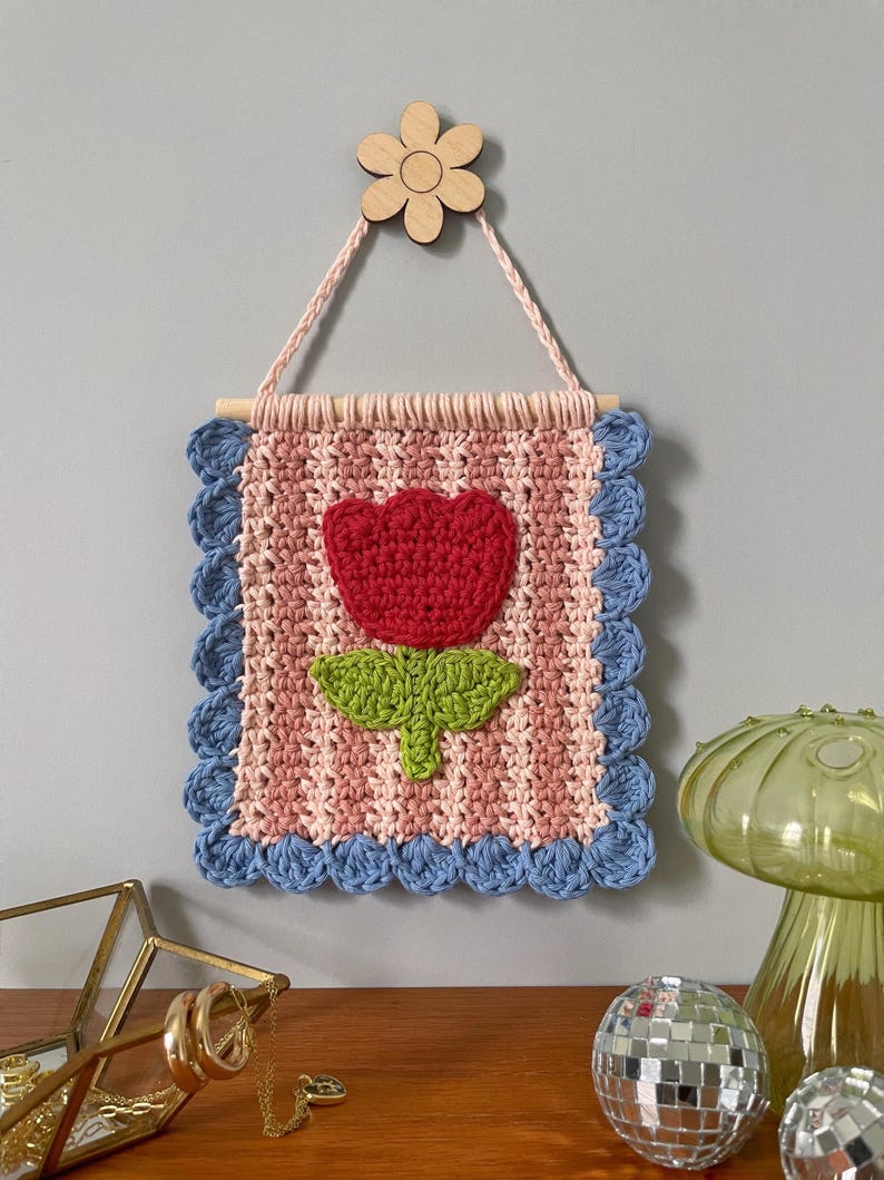 K&ouml;nnte beinhalten: Handgefertigter Wandbehang aus H&auml;kelarbeit mit einer roten Blume und gr&uuml;nen Bl&auml;ttern auf einem rosa-wei&szlig; gestreiften Hintergrund. Die Kanten sind mit blauem H&auml;kelwerk verziert. Eine Holzblume dient zum Aufh&auml;ngen des St&uuml;cks.