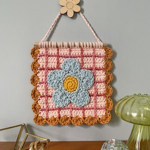 K&ouml;nnte beinhalten: Handgefertigter Wandbehang aus H&auml;kelarbeit mit einer gro&szlig;en blauen Blume mit gelbem Zentrum. Das quadratische Design hat ein rosa-wei&szlig;es Karomuster, das von einem braunen, gewellten Rand umrahmt wird. Eine Holzblume und ein Rundstab zum Aufh&auml;ngen.