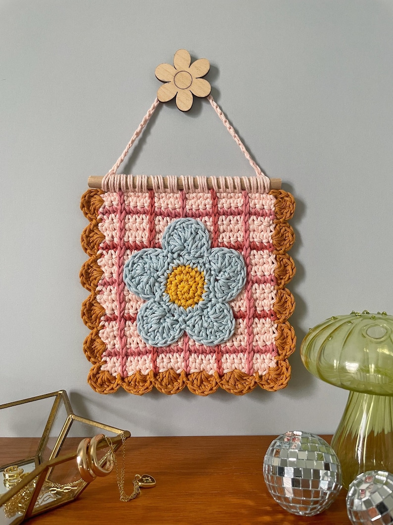 K&ouml;nnte beinhalten: Handgefertigter Wandbehang aus H&auml;kelarbeit mit einer gro&szlig;en blauen Blume mit gelbem Zentrum. Das quadratische Design hat ein rosa-wei&szlig;es Karomuster, das von einem braunen, gewellten Rand umrahmt wird. Eine Holzblume und ein Rundstab zum Aufh&auml;ngen.
