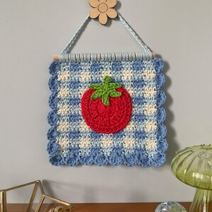 K&ouml;nnte beinhalten: Handgefertigter Wandbehang aus H&auml;kelarbeit mit einer roten Tomate mit gr&uuml;nen Bl&auml;ttern auf einem blau-wei&szlig; karierten Hintergrund. Das quadratische Kunstwerk ist mit einem gewellten Rand versehen und h&auml;ngt an einem Holzstab und einer Holzblume.
