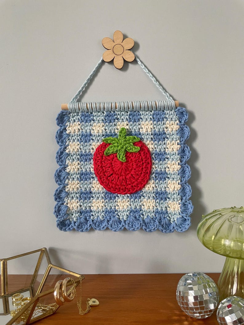K&ouml;nnte beinhalten: Handgefertigter Wandbehang aus H&auml;kelarbeit mit einer roten Tomate mit gr&uuml;nen Bl&auml;ttern auf einem blau-wei&szlig; karierten Hintergrund. Das quadratische Kunstwerk ist mit einem gewellten Rand versehen und h&auml;ngt an einem Holzstab und einer Holzblume.