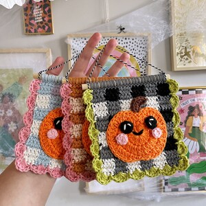 May include: Three handmade crochet pumpkin wall hangings. Each features a smiling orange pumpkin on a checkered background, with scalloped edges in various colors. The hangings are attached to wooden dowels with black and white twine.