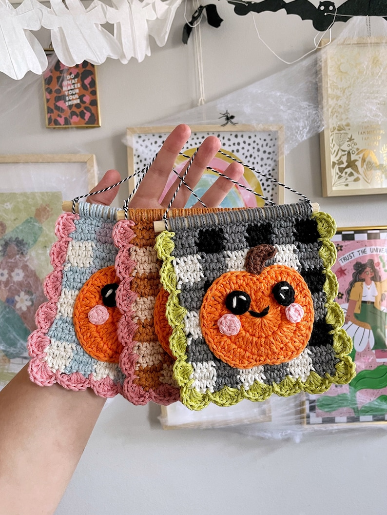May include: Three handmade crochet pumpkin wall hangings. Each features a smiling orange pumpkin on a checkered background, with scalloped edges in various colors. The hangings are attached to wooden dowels with black and white twine.