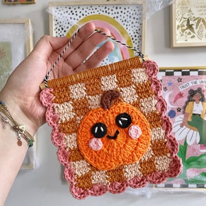 May include: A handmade crochet wall hanging featuring a smiling pumpkin. The pumpkin is orange with black eyes and pink cheeks, set against a brown and cream checkered background. The banner is trimmed with a pink scalloped edge and has a wooden dowel and a black and white string for hanging.