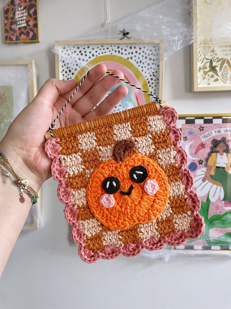 May include: A handmade crochet wall hanging featuring a smiling pumpkin. The pumpkin is orange with black eyes and pink cheeks, set against a brown and cream checkered background. The banner is trimmed with a pink scalloped edge and has a wooden dowel and a black and white string for hanging.
