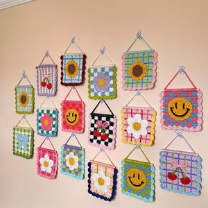 K&ouml;nnte beinhalten: Eine Sammlung farbenfroher, geh&auml;kelter Wandbeh&auml;nge mit verschiedenen Designs, darunter Sonnenblumen, Smiley-Gesichter, Blumen, Kirschen und Schleifen. Jedes quadratische oder rechteckige St&uuml;ck ist mit einem gewellten Rand versehen und wird mit einem kleinen Holzstab und einer Schnur aufgeh&auml;ngt.