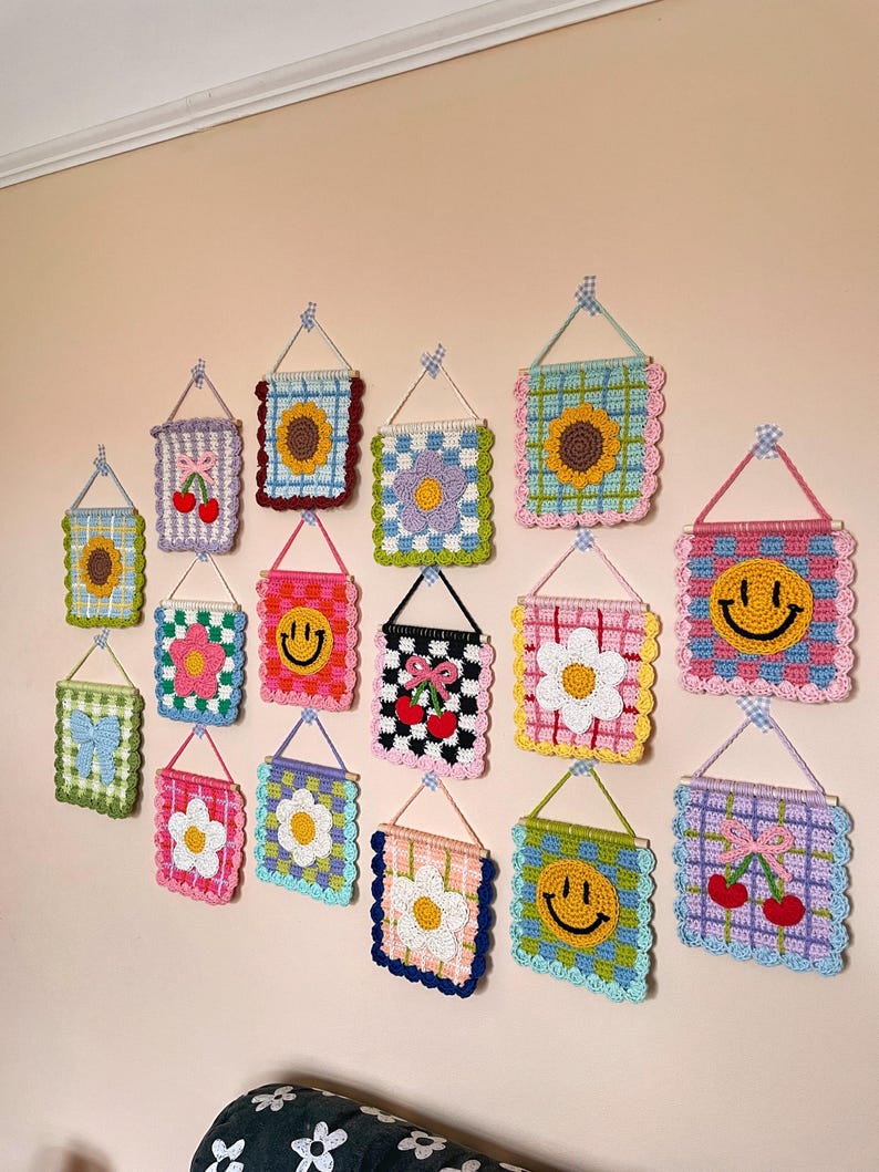 K&ouml;nnte beinhalten: Eine Sammlung farbenfroher, geh&auml;kelter Wandbeh&auml;nge mit verschiedenen Designs, darunter Sonnenblumen, Smiley-Gesichter, Blumen, Kirschen und Schleifen. Jedes quadratische oder rechteckige St&uuml;ck ist mit einem gewellten Rand versehen und wird mit einem kleinen Holzstab und einer Schnur aufgeh&auml;ngt.