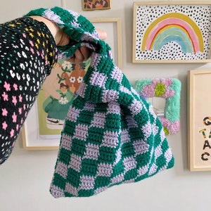May include: A handmade crochet bag in a checkerboard pattern of green and lavender. The bag is held up by a green handle. The bag is a unique accessory.