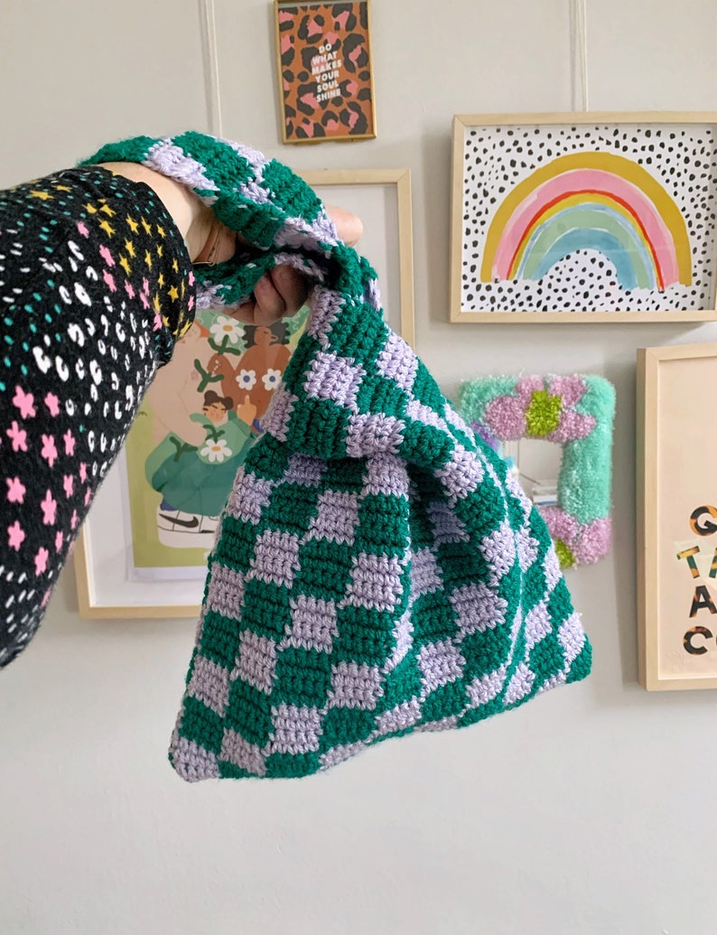 May include: A handmade crochet bag in a checkerboard pattern of green and lavender. The bag is held up by a green handle. The bag is a unique accessory.