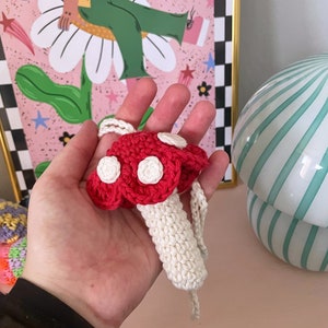 Op de afbeelding: Een gehaakt paddenstoel ornament met een rode dop en witte stippen, en een witte steel. De paddenstoel hangt aan een wit touwtje.