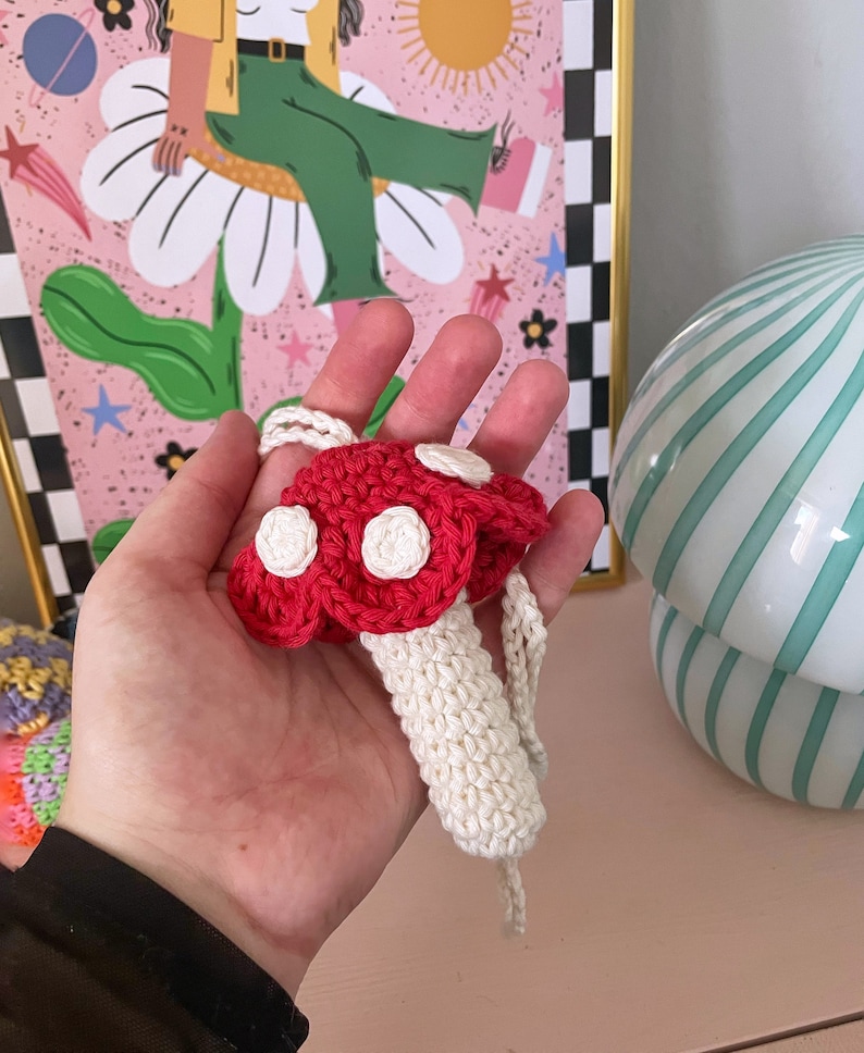 Op de afbeelding: Een gehaakt paddenstoel ornament met een rode dop en witte stippen, en een witte steel. De paddenstoel hangt aan een wit touwtje.