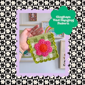 May include: A green, white, and pink gingham crochet wall hanging with a flower design. The hanging is being held by a hand.