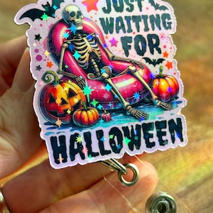 Peut inclure: Un porte-badge à thème Halloween rose et iridescent avec un squelette assis dans une bouée rose gonflable, des citrouilles et des chauves-souris. Le texte indique "Just Waiting For Halloween".