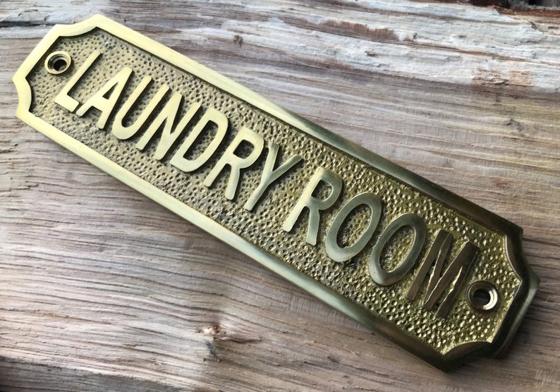 Op de afbeelding: Een messing bord met een gehamerde textuur en afgeronde randen. Het bord heeft de tekst "LAUNDRY ROOM" in reli&euml;f.