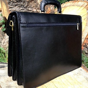 May include: Black leather briefcase with a top handle and a zippered pocket. The briefcase has gold-colored hardware and a structured design. The stitching is visible along the edges, and the briefcase appears to be made of high-quality leather.
