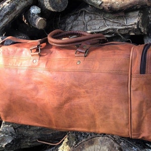 May include: A brown leather duffel bag with two top handles and a zipper closure. The bag is sitting on a pile of logs.