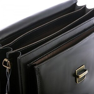 May include: A close-up of a black leather briefcase with multiple interior compartments. The briefcase has a shiny finish and a metal clasp. The interior appears to be lined with a lighter brown material. The briefcase is designed for carrying documents and other professional items.