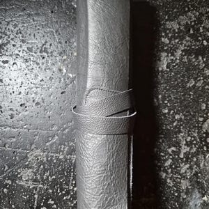 May include: A grey leather pen roll with a textured surface. The roll is secured with a matching leather strap that wraps around the body. The pen roll is designed to hold writing instruments.