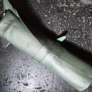 May include: A rolled sage green leather item, possibly a tool roll or organiser, secured with a matching strap. The leather has a textured grain, set against a dark, speckled surface.