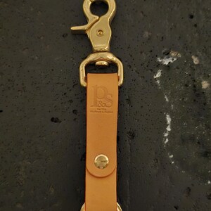 May include: A tan leather keychain with a brass clasp and ring. The leather strap is embossed with the initials "P&S" and the text "Handmade in England". The keychain is designed for attaching keys or other small items.