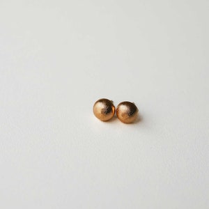 May include: A pair of small, round, gold-colored earrings. The earrings are smooth and have a matte finish.