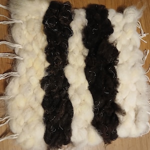 May include: A hand-woven textile featuring alternating stripes of white and dark brown wool. The rectangular piece has white fringe along the edges. The thick, textured wool gives a rustic appearance. The textile is likely a small rug or wall hanging.