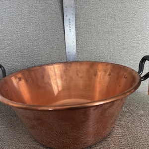 May include: A copper bowl with two black handles. The bowl is round with a wide opening and a hammered texture. A metal ruler is placed inside the bowl. The bowl is sitting on a gray upholstered chair.