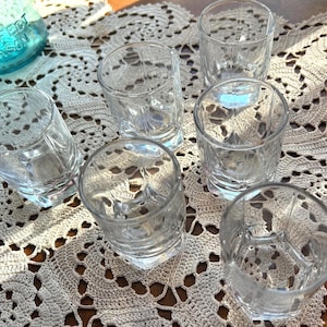 May include: Six clear, patterned shot glasses are arranged on a beige, crocheted doily. The glasses have a geometric design and sit on a wooden surface. A turquoise jar with the text "Perfect Mason" is partially visible.