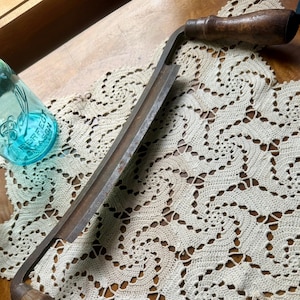 May include: An antique drawknife with a curved blade and wooden handles. The metal blade is a dark gray color, and the handles are a dark brown. The drawknife is resting on a cream-colored, crocheted doily.
