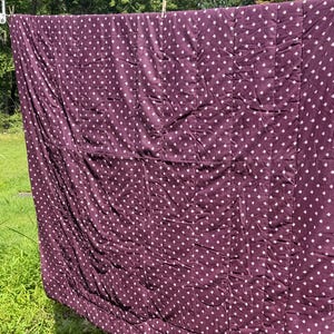 May include: A rectangular quilt featuring a deep plum purple fabric base with a repeating pattern of small white diamond shapes. The quilt is constructed with vertical and horizontal stitched seams and is suspended from a clothesline outdoors.