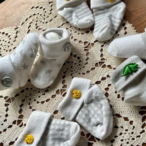 May include: A set of ten baby socks with different designs. The socks are white with grey stripes and feature various buttons, including a yellow duck, a green dinosaur, and a white snowflake.