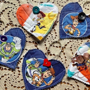 May include: Five handmade heart-shaped ornaments featuring characters from the Toy Story franchise. The ornaments are made of fabric and feature different characters, including Woody, Buzz Lightyear, and Jessie. Each ornament has a button sewn on the front.
