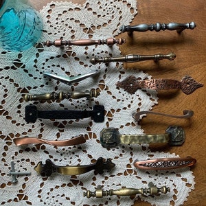 May include: Assortment of vintage cabinet hardware in various styles and finishes, including copper, brass, and black. The hardware is arranged on a white crocheted doily.