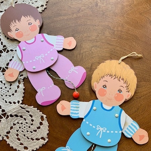 May include: Two wooden pull string puppets, one with a pink dress and the other with a blue dress. Both puppets have brown hair and painted faces.