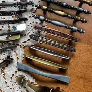 May include: Assortment of decorative cabinet hardware in various styles and finishes, including brass, copper, and silver. The hardware features a variety of designs, including ornate, geometric, and simple.