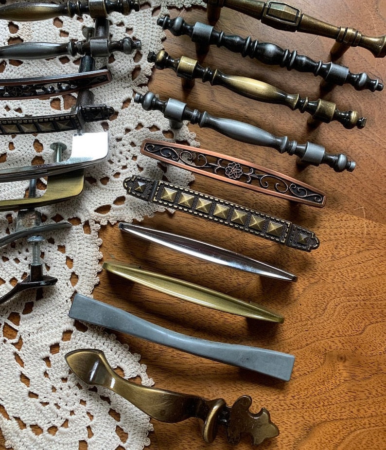 May include: Assortment of decorative cabinet hardware in various styles and finishes, including brass, copper, and silver. The hardware features a variety of designs, including ornate, geometric, and simple.