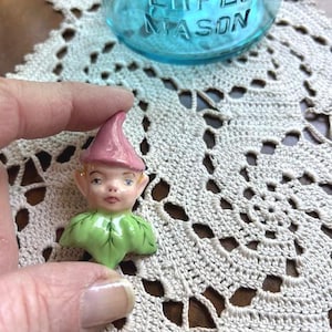 May include: A small ceramic figurine head featuring a pointed pink hat and a green leaf-shaped collar. The face has painted blue eyes, rosy cheeks, and a neutral expression. The piece is crafted with a glossy finish and detailed facial features.