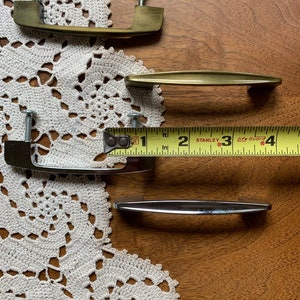 May include: Four cabinet pulls, two are gold and two are silver. The gold pulls are a simple, curved design. The silver pulls are a more modern, straight design. A measuring tape is in the image, with the numbers 1, 2, 3, and 4 visible.