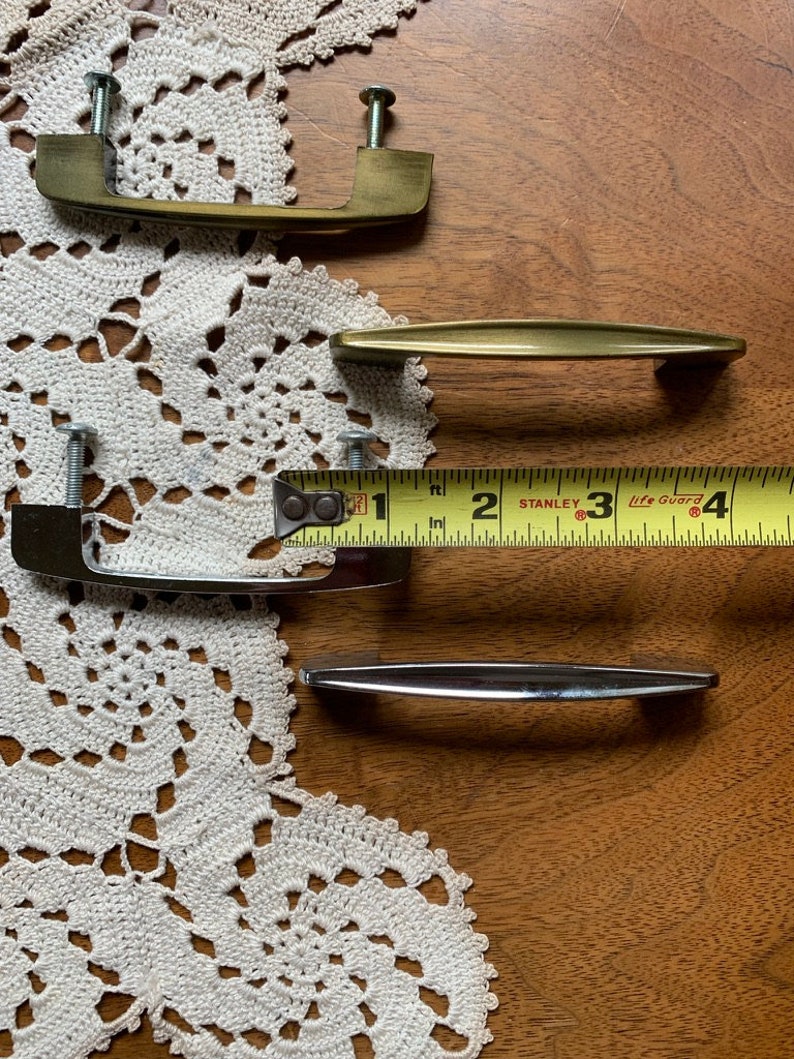 May include: Four cabinet pulls, two are gold and two are silver. The gold pulls are a simple, curved design. The silver pulls are a more modern, straight design. A measuring tape is in the image, with the numbers 1, 2, 3, and 4 visible.