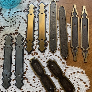 May include: A collection of eight antique-style metal drawer pulls in various finishes, including brass, silver, and black. The pulls have ornate designs and are arranged on a white crocheted doily.