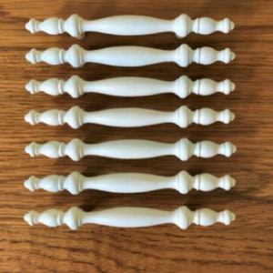 May include: Set of seven white cabinet knobs with a decorative, slightly curved design. The knobs have a smooth, matte finish.